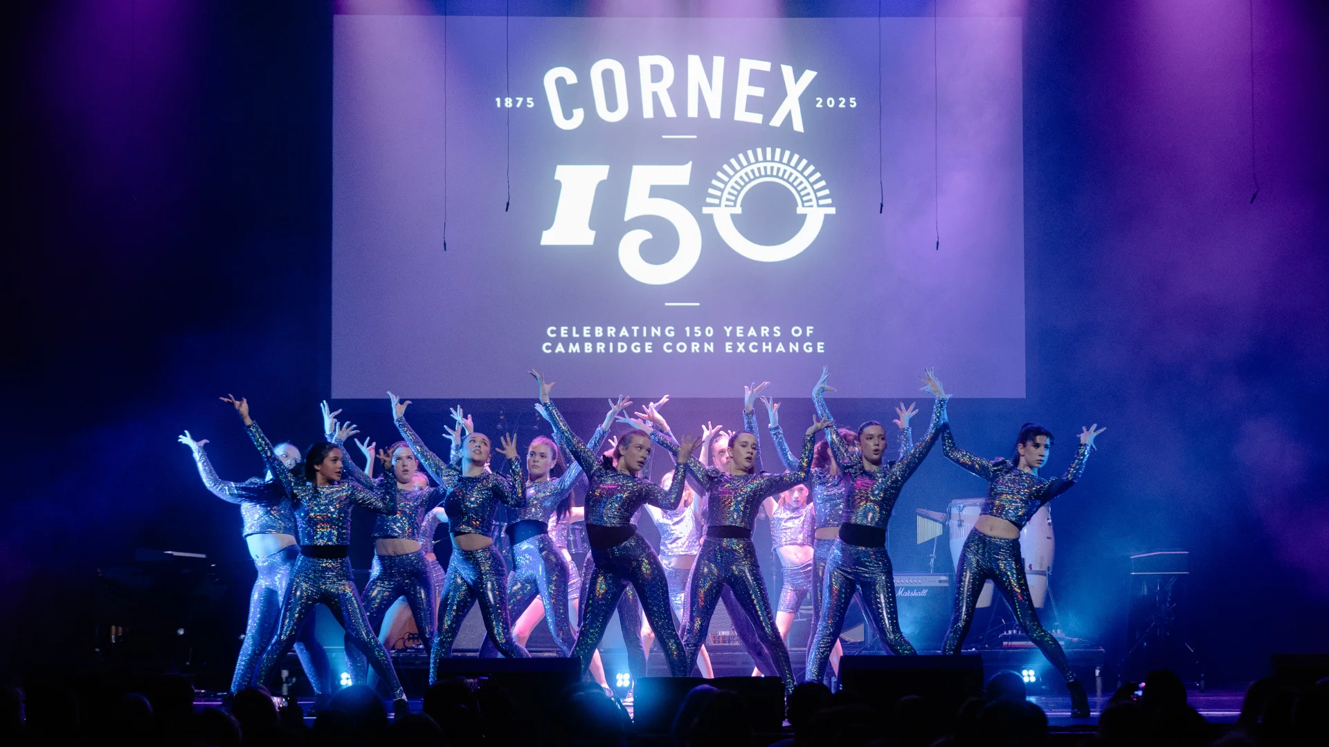 Image shows a coloured photograph of a troop of young female dancers dressed in metallic costumes performing on the Cambridge Corn Exchange stage in front of 150 years of Corn Exchange backdrop