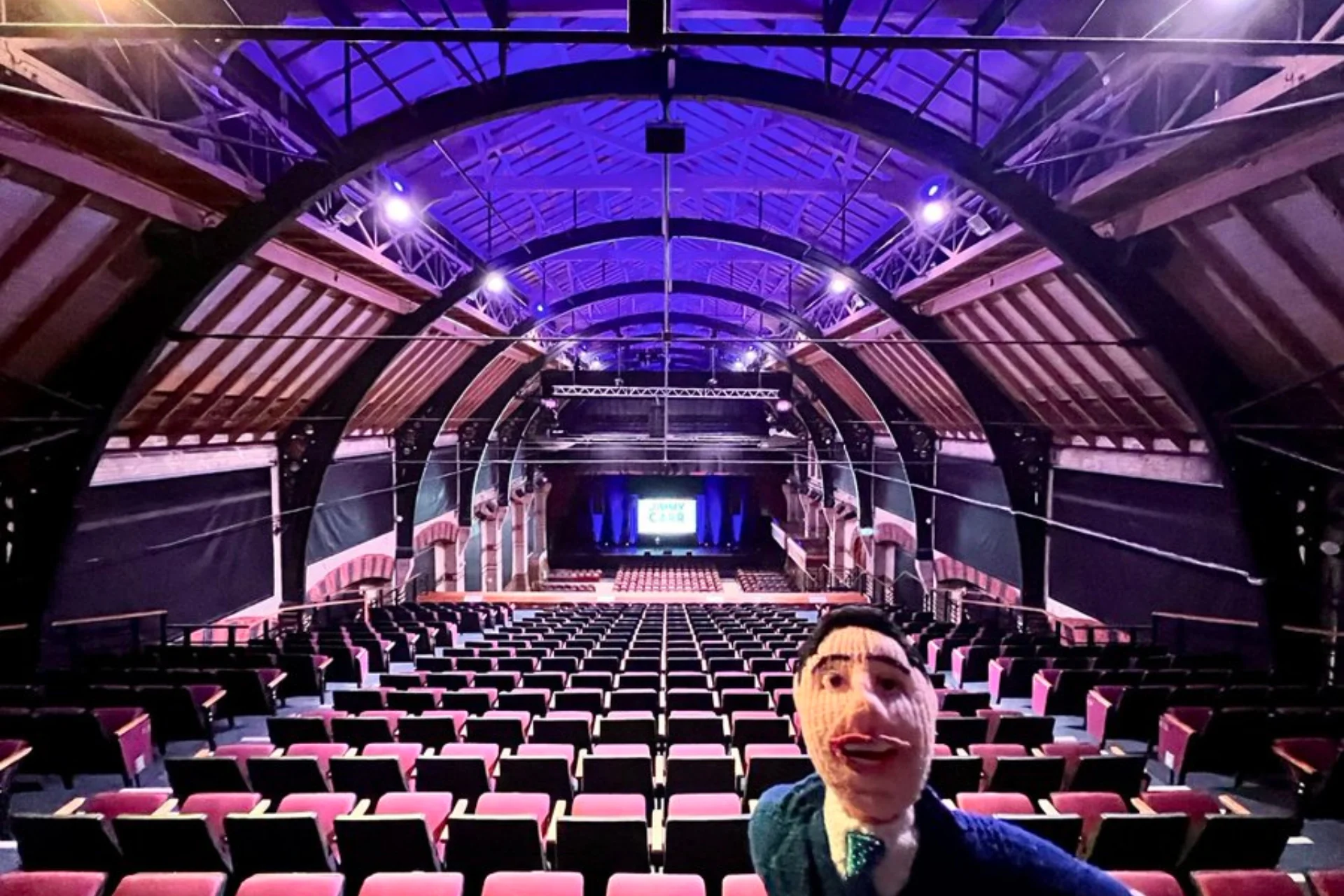 Photographic image shows the interior of the Cambridge Corn Exchange taken from the back of the venue facing the stage. In the foreground of the picture is a knitted puppet.