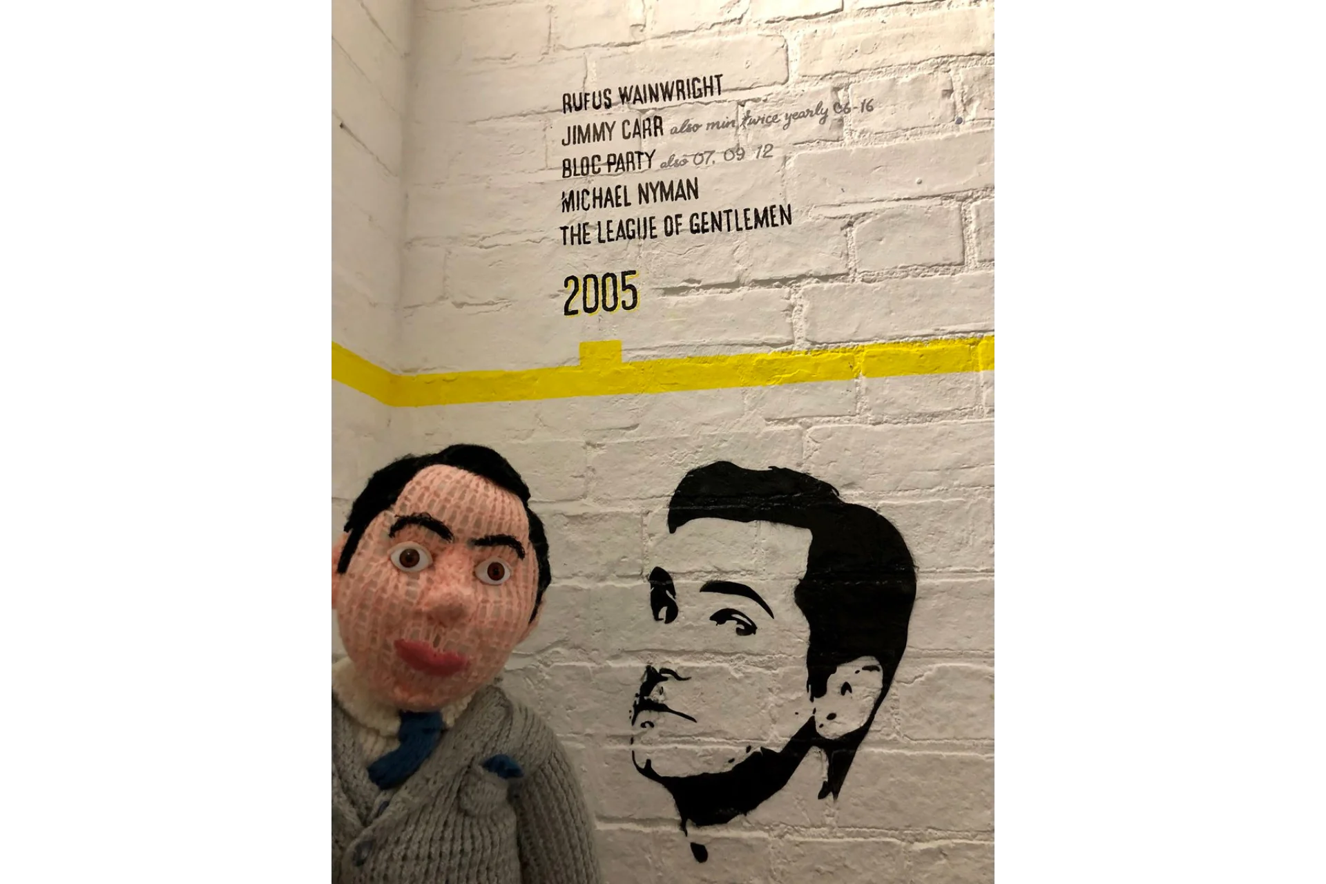 Photographic image of a knitted puppet that resembles comedian Jimmy Carr in front of the Corn Exchange painted timeline mural that is yellow and painted on a white brick wall. There is a stencilled black spray painted image of comedian Jimmy Carr as part of this timeline along with his name listed under 2005 along with other performers.