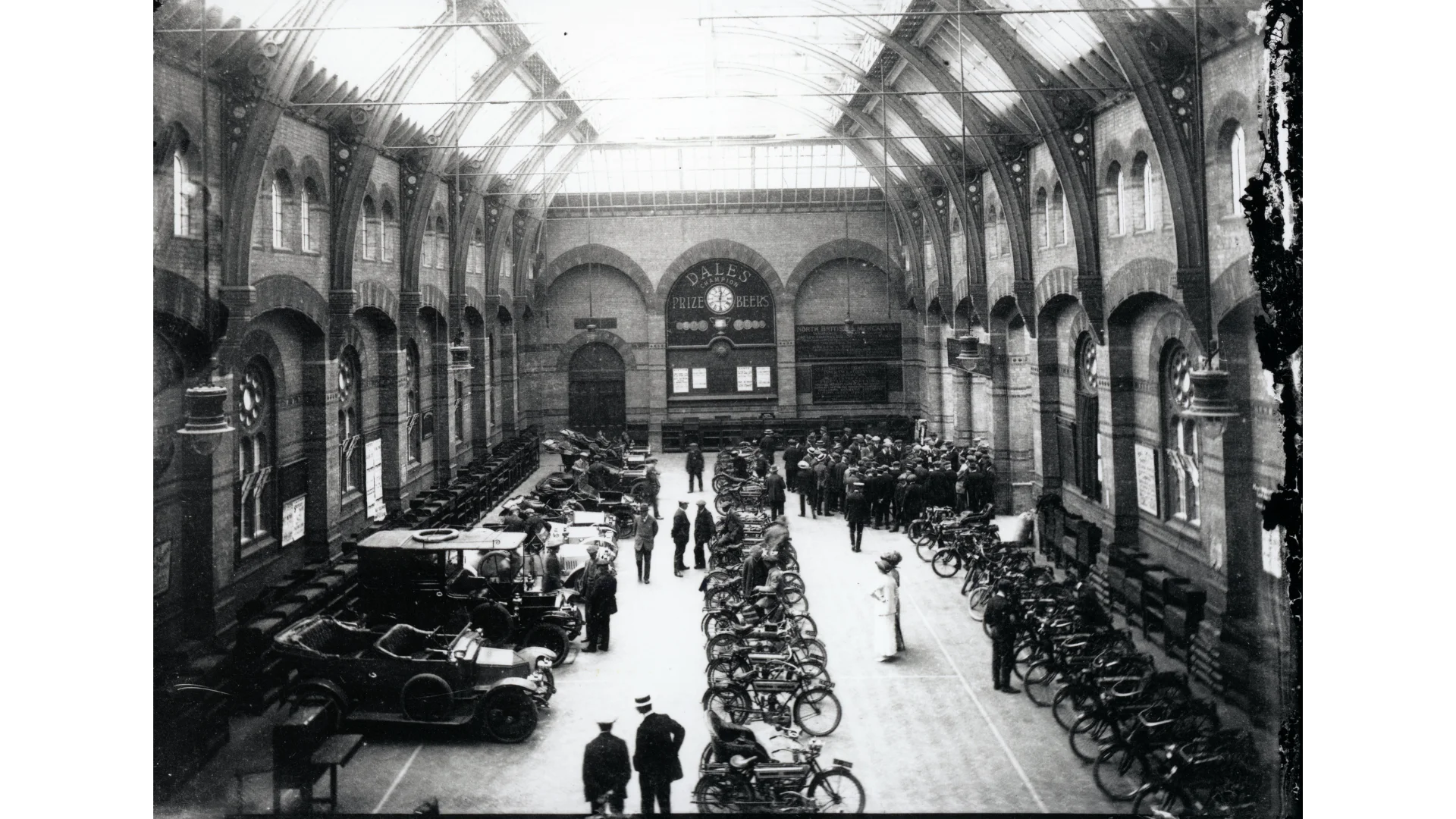 An old black and white photograph of the Corn Exchange when it had a glass ceiling. Old vintage cars fill the hall as well as bikes lined up.
