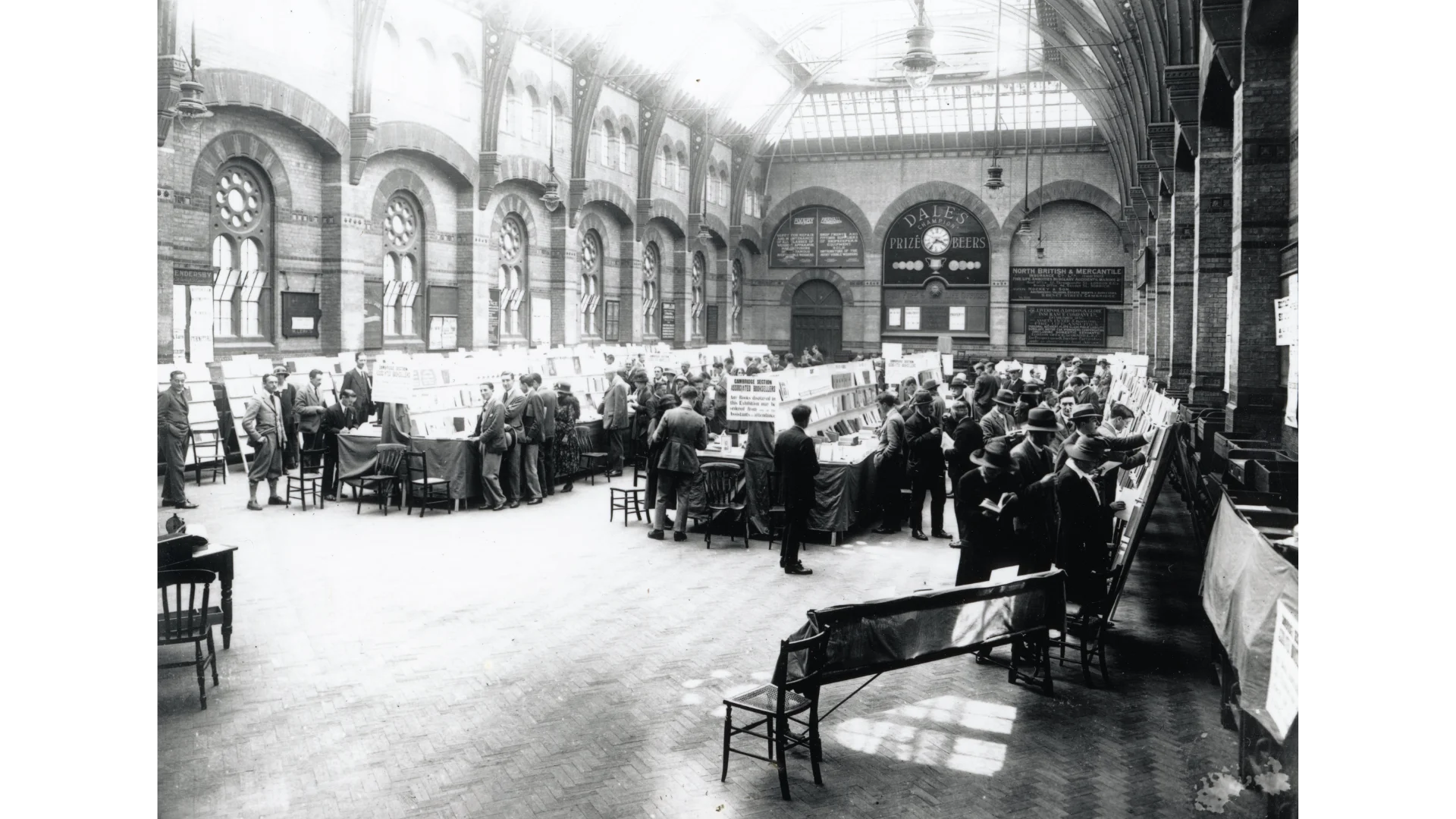 An old black and white photograph of the Corn Exchange when it had a glass ceiling, with rows of stalls in the hall selling books. There are crowds of people in suits browsing.