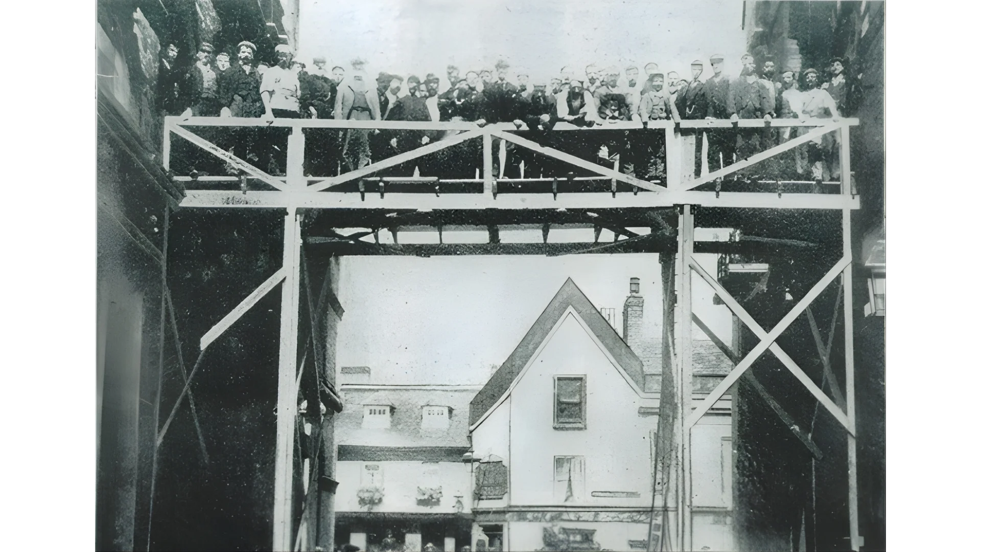 An old black and white image of a scaffolding-like bridge between two buildings. There are over 40 people stood on top of it looking down at the camera.
