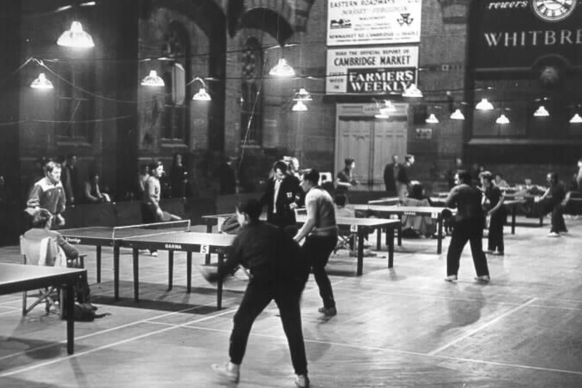 An old black and white photograph inside the Corn Exchange. The hall is filled with table tennis players, and there is low strung lighting highlighting the tables.