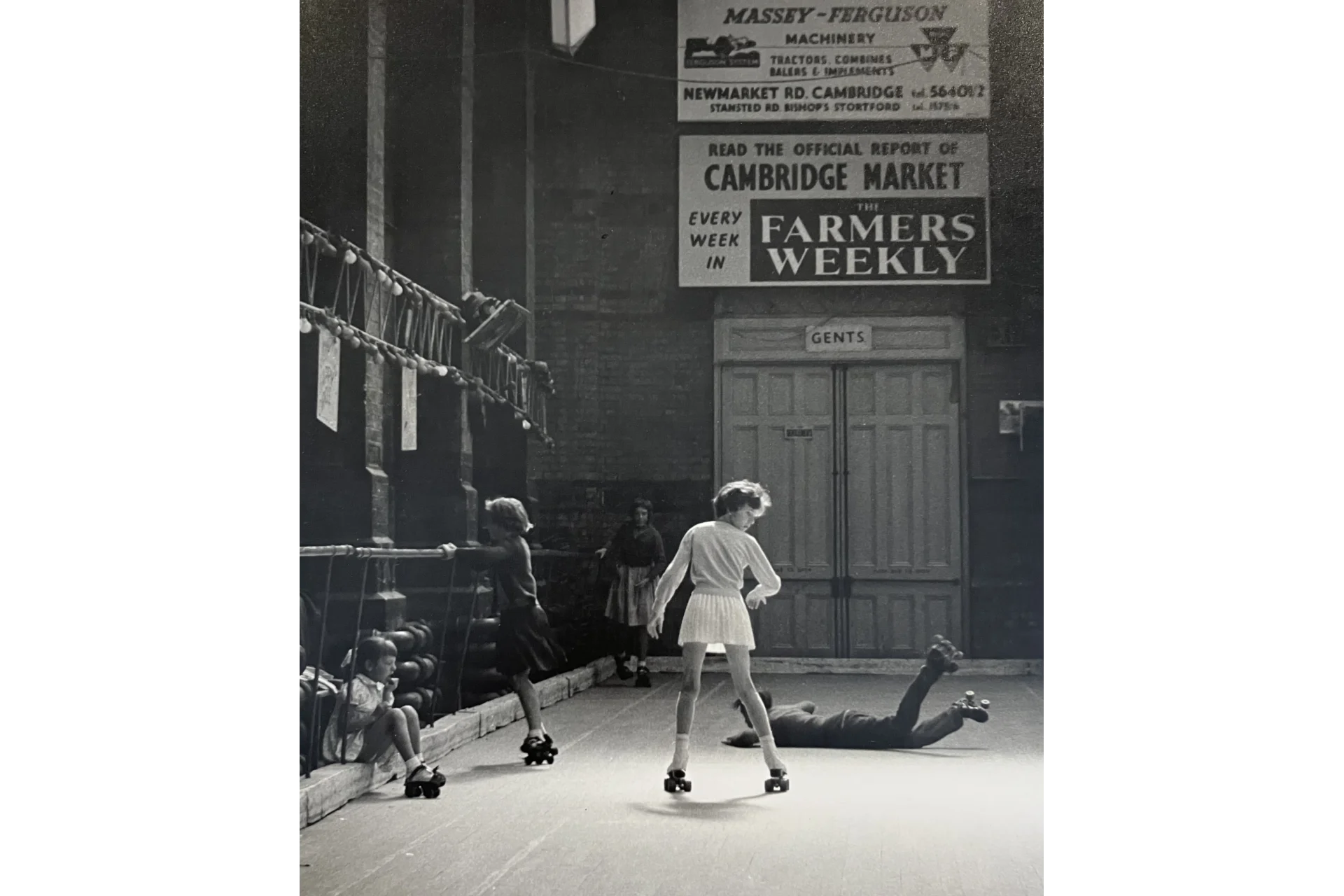 An old black and white photograph of children roller-skating inside the Corn Exchange. There are two children sat and leaning on the banister, and a girl in a white top and skirt is in the centre. A child lays on the floor beside her. There are advertisements for local businesses above the door.