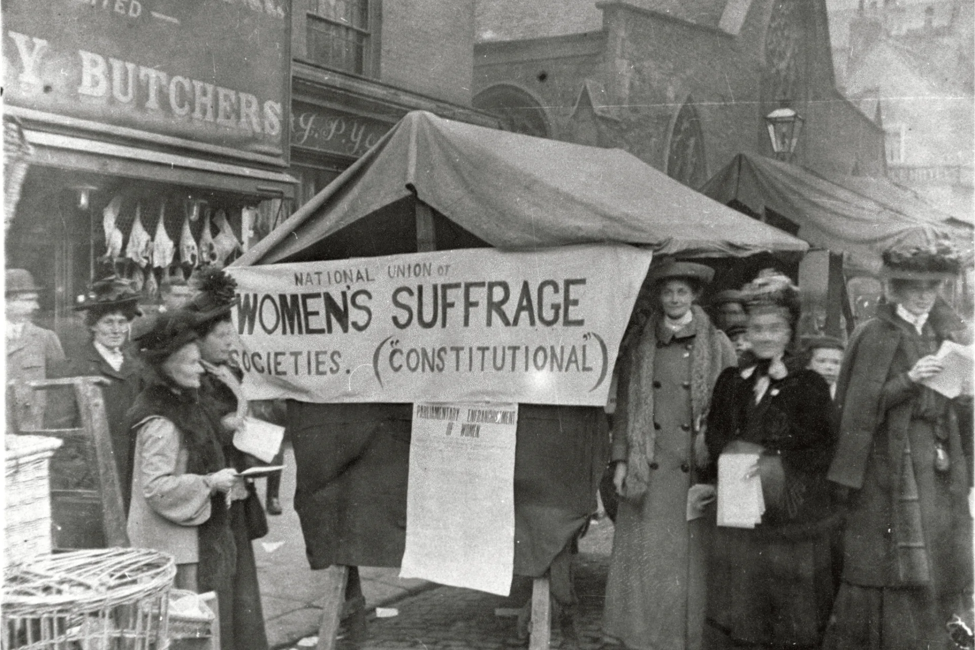 Image shows a black and white photographic print of a suffrage protest in market square. The image is blurred at the edges and shows six women dressed in Edwardian era full length coats and elaborate feathered hats with young children standing behind them. A clothed banner that reads National Union of Women's Suffrage hangs on a stall in the centre of the group.