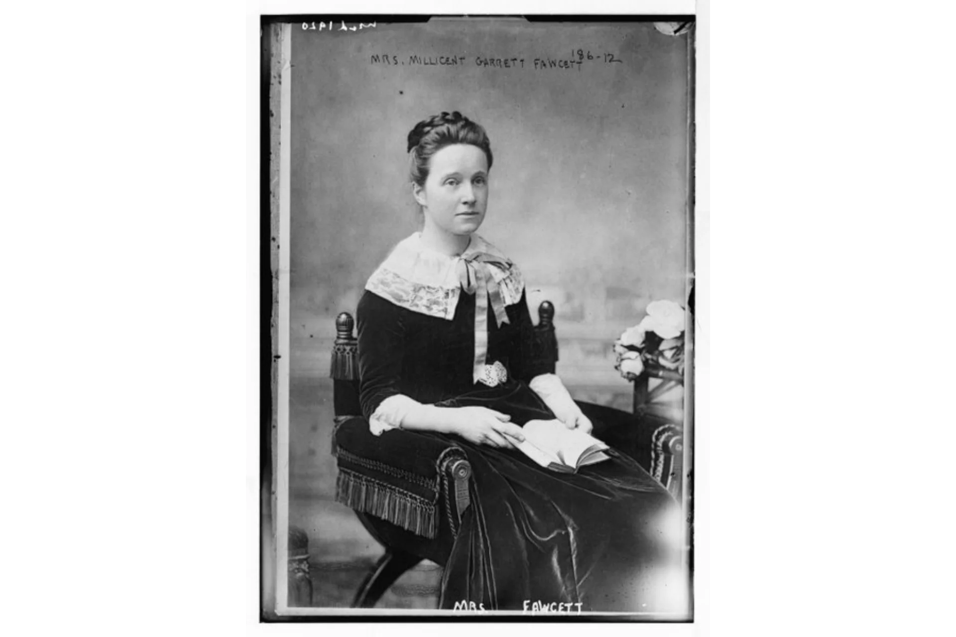 Image shows a black and white photographic print portrait of Millicent Fawcett sitting in a chair as if on a throne. She is wearing Edwardian velvet dress and is holding an open book on her lap.